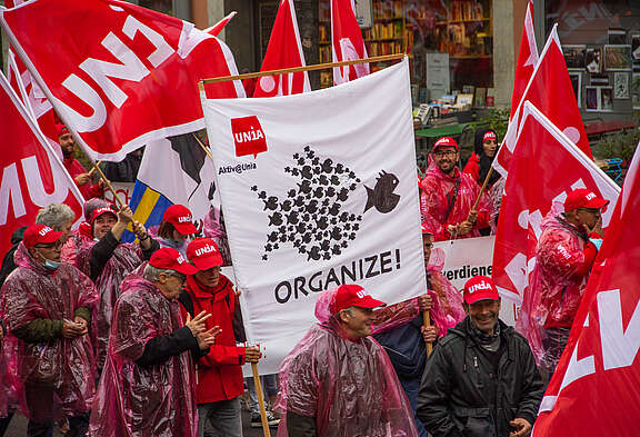 Demonstrierende mit roten Unia-Fahnen und Transparent «ORGANIZE!» fordern bessere Arbeitsbedingungen. Viele tragen rote Kappen und Regencapes.