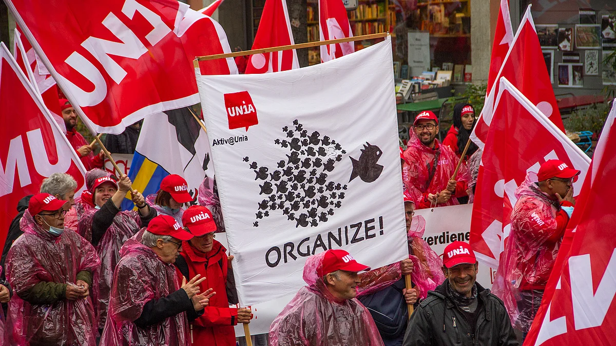 Demonstrierende mit roten Unia-Fahnen und Transparent «ORGANIZE!» fordern bessere Arbeitsbedingungen. Viele tragen rote Kappen und Regencapes.